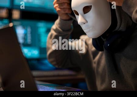 Closeup of person wearing anonymity mask works on laptop, representing online security threat or hidden identity theme. Masked male individual wearing hoodie in tech room lit by multiple data screens. Stock Photo
