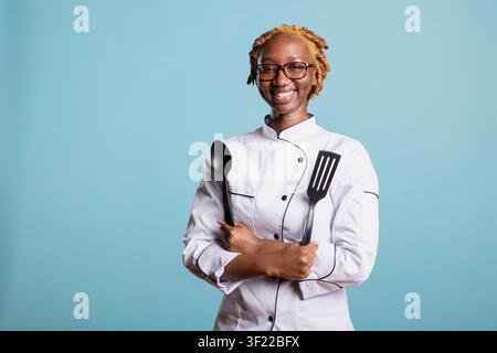 Cheerful female chef posing with crossed arms, holding plastic spatula and spoon in studio. African american food expert smiles warmly, showing passion for cooking and friendly culinary personality. Stock Photo