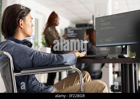 Asian programmer in wheelchair typing code at inclusive tech office desk, focused on software development and accessibility solutions. Disabled developer analyzes backend data with concentration. Stock Photo