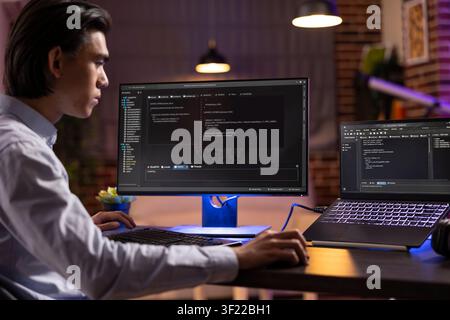 Self employed man sits at desk surrounded with computers, reviewing software and analyzing website structure. Young male adult working from home, completing programming tasks during late hours. Stock Photo