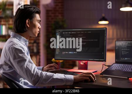 Young male entrepreneur spending late evening hours at home improving app performance and functionality. Focused programmer working from home and analyzing html code displayed across computer screens. Stock Photo