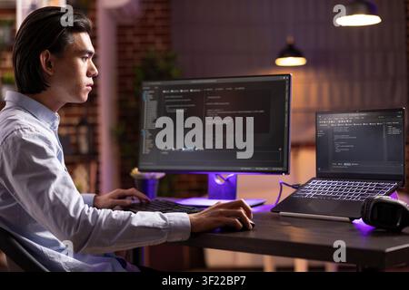 Remote asian worker sits with multiple screens coding online applications and debugging website errors. Male software developer enjoys flexibility of working from home and digital entrepreneurship. Stock Photo