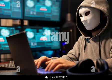 Masked hacker attempts data breach, targeting sensitive digital information in tech environment. Freelance programmer anonymously writes malware code on laptop, hacking into system software. Stock Photo