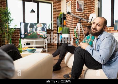 Bearded white man plays video game in living room as black friend enjoys beer nearby. Caucasian male individual talking with group while gaming on TV console, sharing fun and leisure at home. Stock Photo