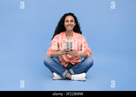 Woman Sits Cross-Legged on Blue Background While Using a Smartphone to Connect With Others Stock Photo