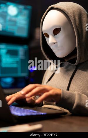 Anonymous programmer conceals identity while typing malicious code on laptop. Hacker works late at night and targets company networks, symbolizing cybersecurity threats, online crime and data breaches Stock Photo