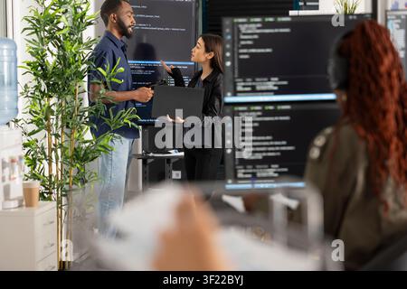 Male and female software developers stand by computer monitors, brainstorming coding project for startup. Diverse team collaborates on problem solving HTML scripts while building and refining website. Stock Photo