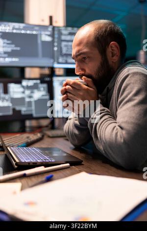 Caucasian male programmer clasping hot drink to stay awake and reviewing error messages on screen. Bearded man looks anxious, working night hours in cybersecurity environment full of active monitors. Stock Photo