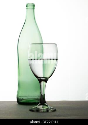 A minimalist still life composition featuring an empty green glass bottle standing behind a clear wine glass filled with water. Stock Photo