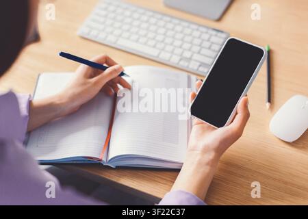 Unrecognizable Person Writing Notes In Journal While Holding Smartphone, Office Setting Stock Photo