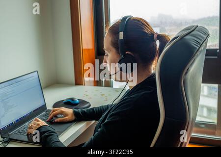 Woman wearing headset with microphone, working remotely on laptop from home office Stock Photo