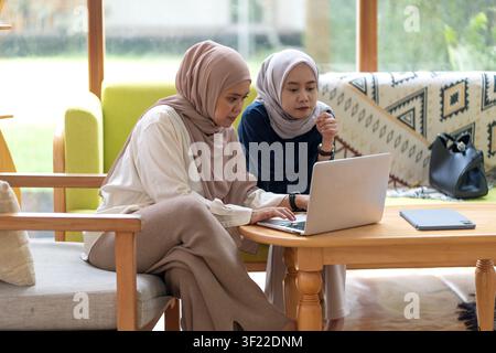 Two Indonesian Muslim professionals working together on a laptop, discussing ideas and collaborating in a comfortable modern workspace. Stock Photo