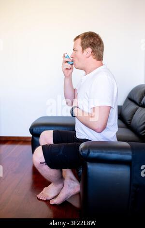Man suffering from asthma sitting on a sofa and using an inhaler for breathing relief Stock Photo