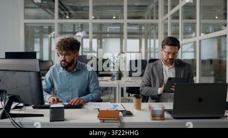 Two multiracial business men working in office with computers Arabian intern businessman worker has problem difficulty ask help Caucasian male boss Stock Photo