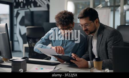 Two multiracial business men working with documents computer talking holding paper checklist analyzing chart graph businessman talk efficiency of job Stock Photo