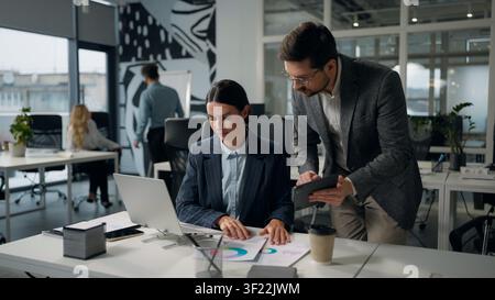 Two Caucasian business colleagues talking discuss data in digital tablet compare information in laptop businesswoman female with businessman male Stock Photo