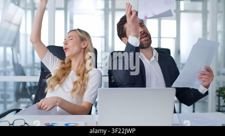 Two happy active business colleagues throwing papers singing dancing celebrating complete paperwork feeling free at office table Caucasian businessman Stock Photo