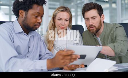 Happy multiethnic business team looking in digital tablet app smiling laughing talking three business colleagues using portable computer with Stock Photo