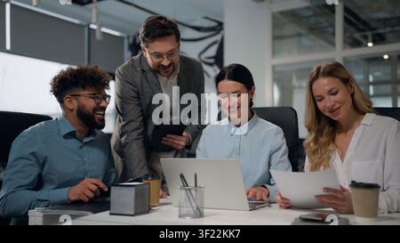 Four laughing happy diversity business workers fun at conference briefing teamwork in office talking looking in laptop analyzing documents laugh Stock Photo