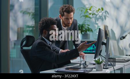 Two multiethnic business men working in office. African American leader work with computer talking advise to Caucasian manager asking help with Stock Photo