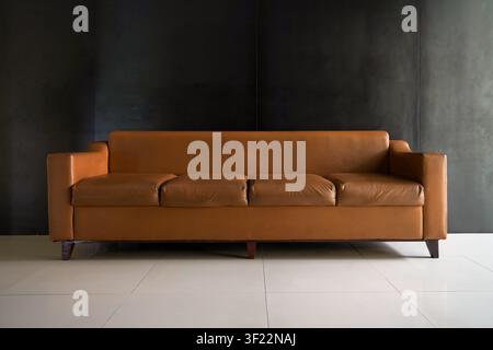 Stylish brown leather sofa with clean lines set against a dark textured wall and white tile floor in a minimalist interior space Stock Photo