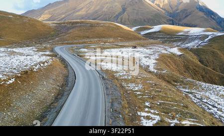 Horizontal high angle view of an empty curved highway stretching through rolling hills with patches of first winter snow under Caucasus mountains in G Stock Photo