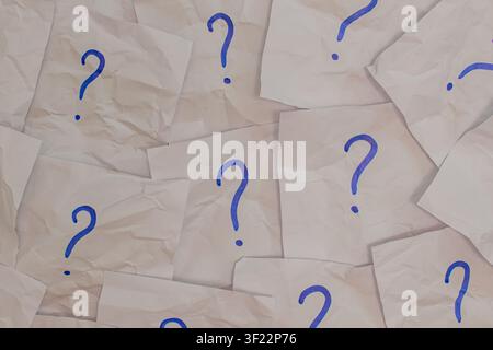 Horizontal top down view of many crumpled white paper notes with blue handwritten question marks scattered on a flat surface representing confusion an Stock Photo
