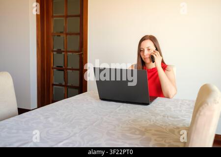 Woman working remotely at home with laptop on table and communicating using phone Stock Photo