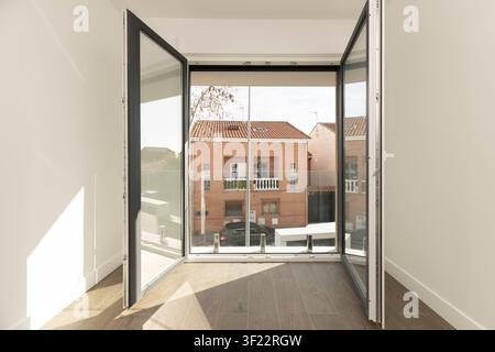 Sunlit hallway framed by open glass balcony doors, overlooking brick townhouses, terracotta rooftops, and parked cars on residential street below. Stock Photo