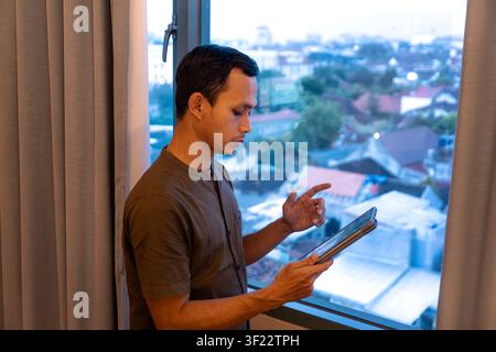 Young asian man standing by a window while using a tablet, focused and engaged, with urban cityscape outside the window. Stock Photo
