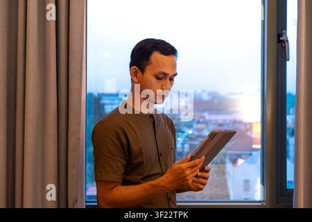 Young asian man standing by a window while using a tablet, focused and engaged, with urban cityscape outside the window. Stock Photo