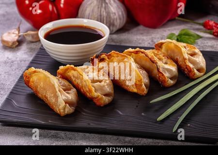 Fried dumplings with golden brown crust arranged on black plate with soy sauce bowl. Red peppers, garlic, green onions in background. Warm lighting hi Stock Photo