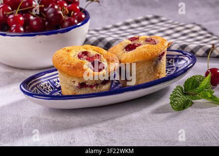Golden cherry muffins on blue white ceramic plate with fresh cherries mint. Soft natural lighting highlights textures. Rustic checkered cloth backgrou Stock Photo