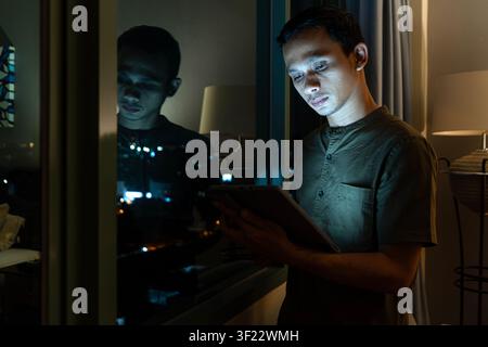 Focused asian young man using a tablet by the window at night, illuminated by screen glow and city lights reflection. Stock Photo