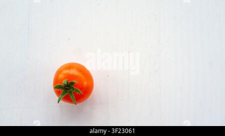 Single ripe red tomato with green calyx rests on pale wooden background, seen from overhead in bright natural light, offering fresh food styling, simp Stock Photo