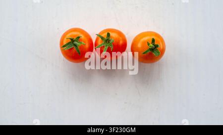 Three ripe cherry tomatoes with green stems arranged in neat row on clean white background, fresh red produce in overhead view with minimal compositio Stock Photo