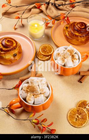 Warm hot chocolate with marshmallows and cinnamon dusting in pumpkin shaped mugs with cinnamon rolls, dried orange slices, a lit candle, and autumn de Stock Photo