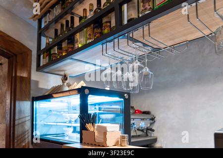 A kitchen shelf contains various bottles of oil and spices. Below, there are hanging glasses and a holder with utensils. This setup is in a restaurant Stock Photo
