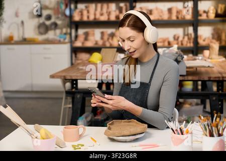Beautiful woman potter in an apron using her phone while working on clay pots Stock Photo
