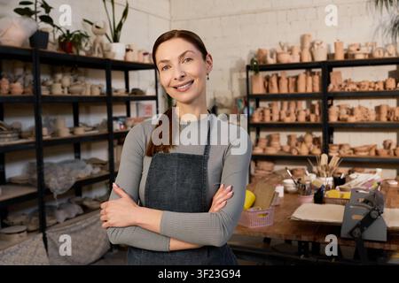 A skilled woman wearing an apron smiles while shaping clay in her warm studio Stock Photo