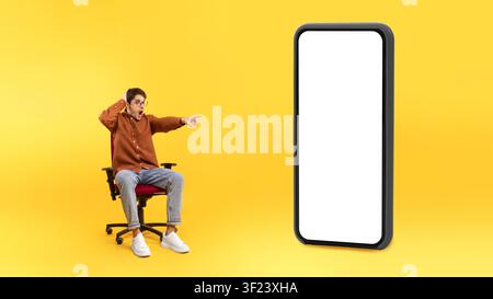 Surprised Young Male Pointing At Blank Smartphone Screen, Yellow Background, Mockup Stock Photo