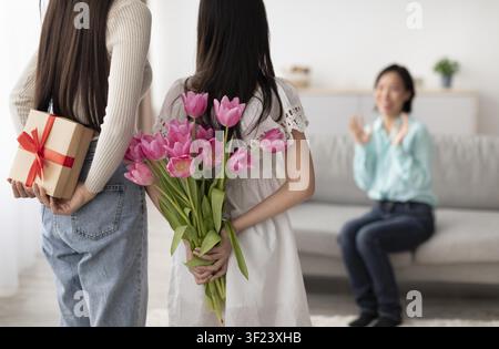 Two Young Women Holding Flowers And Gift Surprise For Female Friend At Home, Celebration Atmosphere Stock Photo