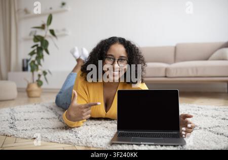 Smiling African American Woman Pointing At Laptop Screen While Lying On Carpet At Home Stock Photo