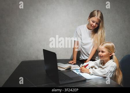 Mother helping her daughter with online school education at home, child reading writing and studying using laptop homeschooling digital distance learn Stock Photo