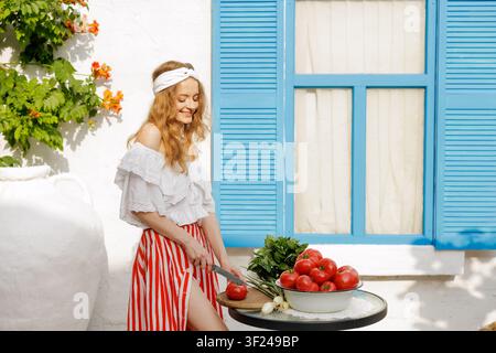 A joyful woman slices ripe tomatoes in a sunny outdoor kitchen, preparing fresh ingredients for gazpacho and salsa, surrounded by vibrant greenery. Stock Photo