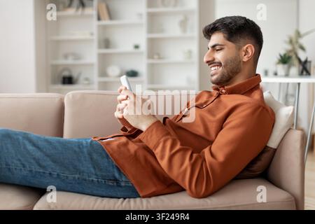 Smiling Young Asian Man Lying On Couch Using Smartphone At Home, Relaxing Lifestyle Concept Stock Photo