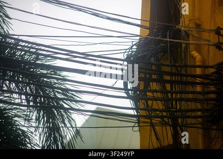 A cluster of tangled electrical wires and cables is attached to a utility pole next to a building, with a cloudy sky in the background and some tree f Stock Photo