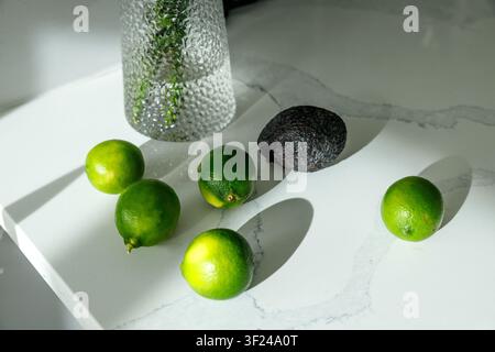 Five bright green limes and a ripe avocado rest in warm directional sunlight on a white marble countertop beside a textured glass vase, casting long s Stock Photo