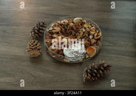 A glass bowl of dried botanical potpourri with a white ornamental stone sits among three pine cones on a dark wooden floor, a warm rustic arrangement Stock Photo