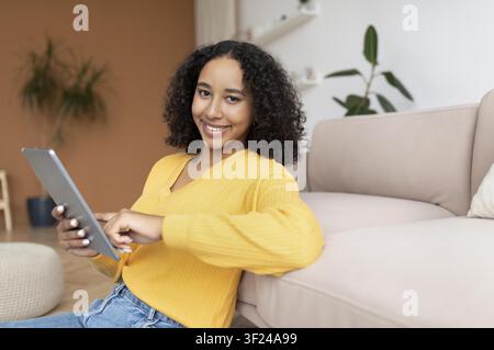 Smiling Young Black Woman Using Tablet At Home, Relaxed Living Room Setting Stock Photo